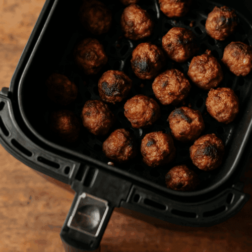 Geöffneter schwarzer Airfryer-Korb gefüllt mit goldbraunen Mini-Frikadellen, Blick von schräg oben auf Holztisch