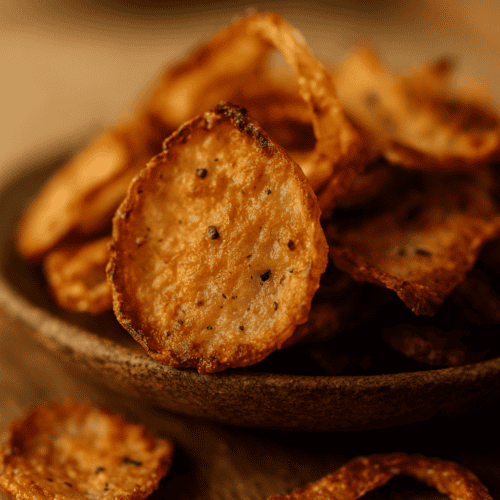 Knusprige goldbraune Zwiebel-Chips in rustikaler Holzschale mit schwarzem Pfeffer auf Holztisch