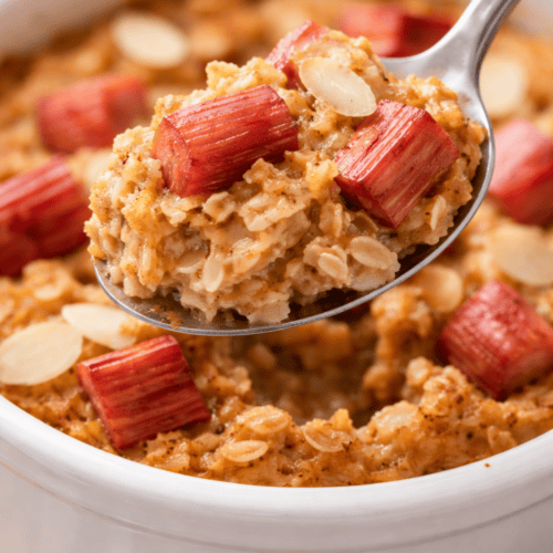 Löffel hebt einen Bissen Rhabarber Baked Oats aus der weißen Auflaufform – cremige Haferflocken mit Rhabarberstücken