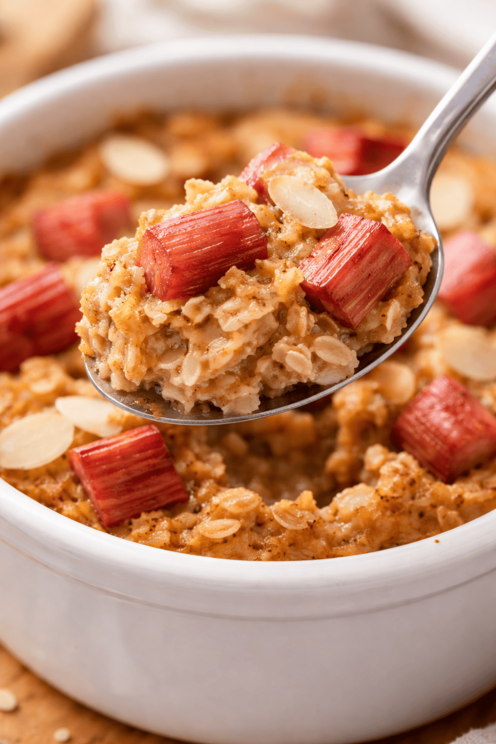 Löffel hebt einen Bissen Rhabarber Baked Oats aus der weißen Auflaufform – cremige Haferflocken mit Rhabarberstücken