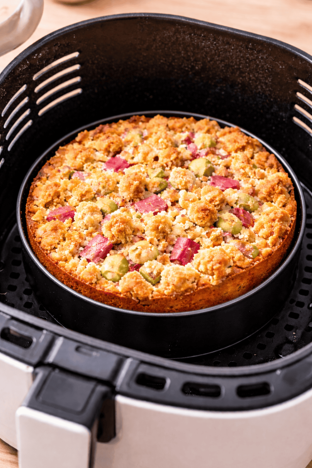 Rhabarber-Streuselkuchen in einer runden Backform Ø 18 cm im Korb einer weißen Heißluftfritteuse – fertig gebacken mit knusprigen Streuseln.