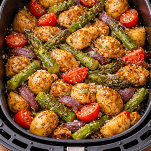 Spargelpfanne im Airfryer-Korb mit grünem Spargel, Hähnchenstücken, vorgegarten Kartoffeln, roten Zwiebeln und halbierten Tomaten, bereit zum Garen